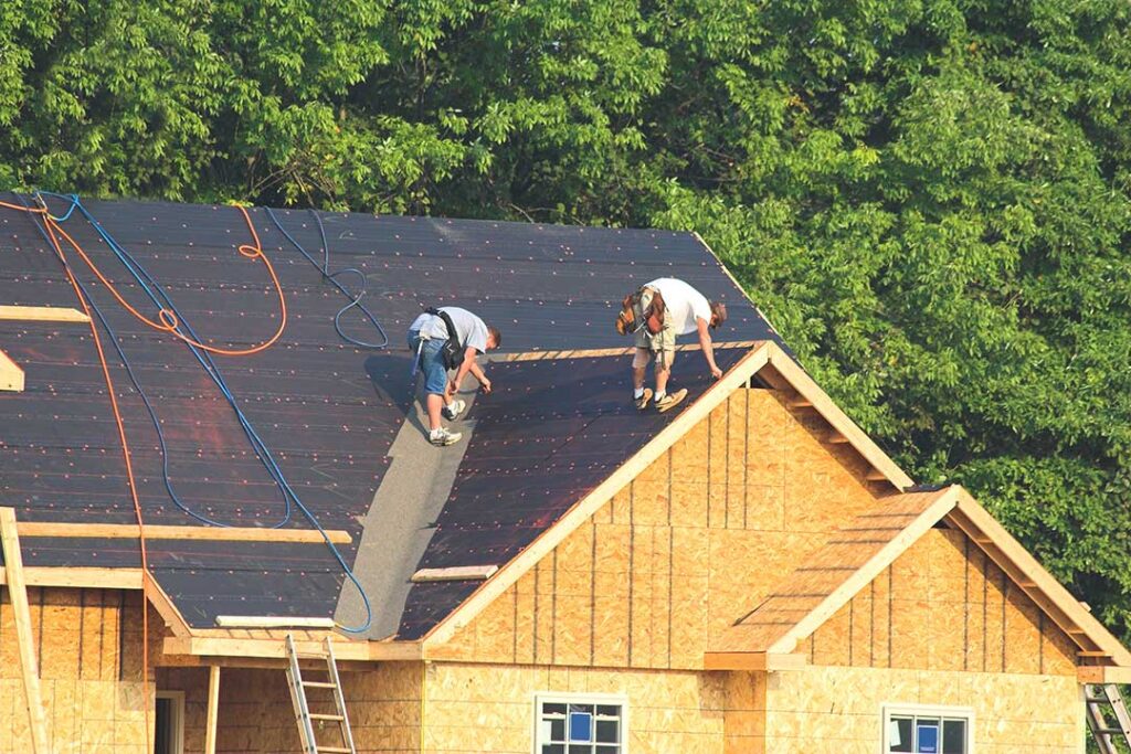 A snow-dusted residential roof with reflective shingles, solar sections, and a technician inspecting layered roofing materials.