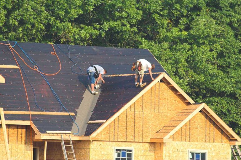 A snow-dusted residential roof with reflective shingles, solar sections, and a technician inspecting layered roofing materials.